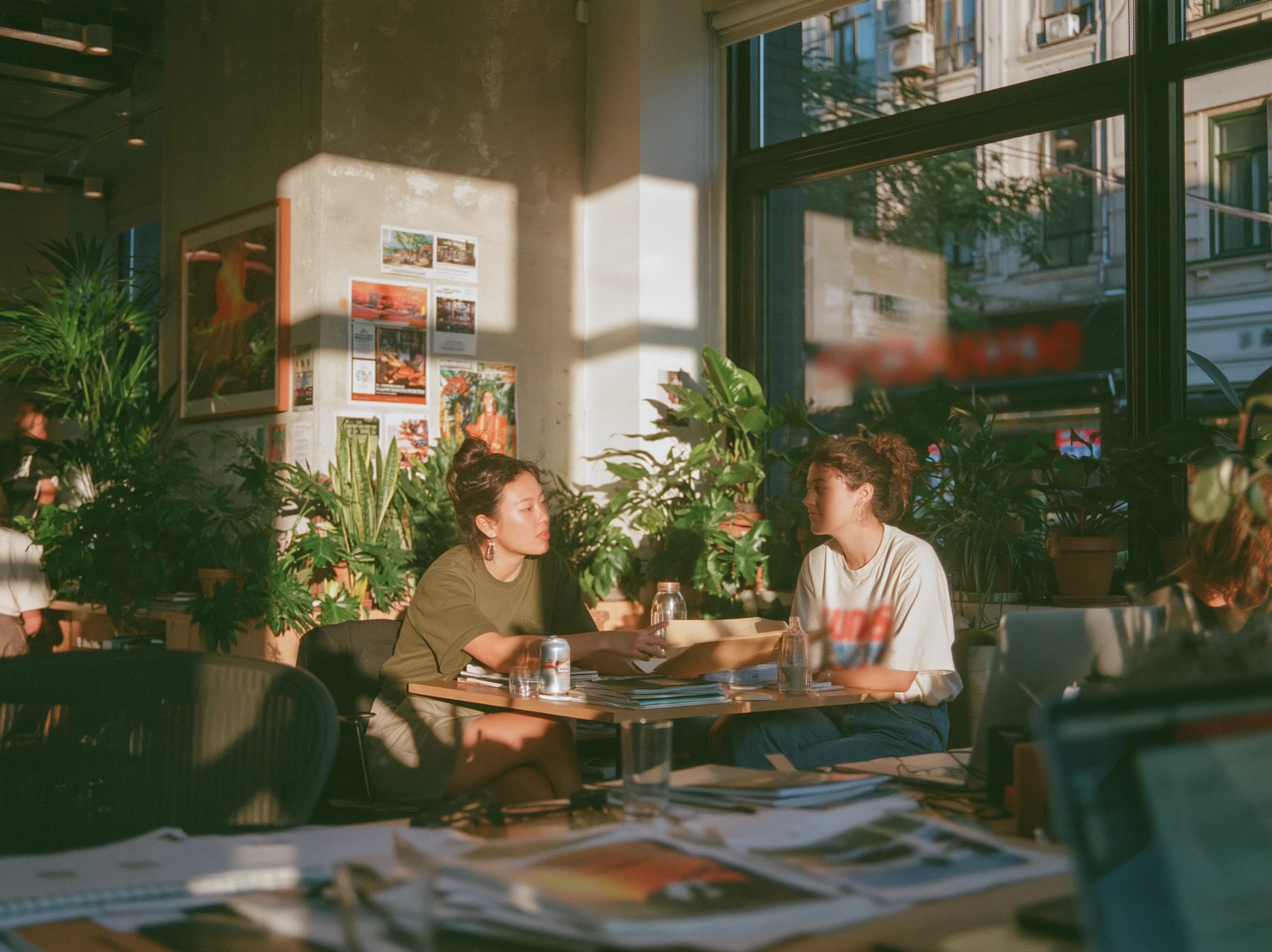 Two women at a small table in a sunny office full of plants, one sliding a folder across to the other.